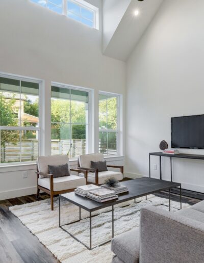 Modern living room with natural light, featuring a gray sofa, coffee table, chairs, and a television, with a view of the backyard through sliding glass doors.