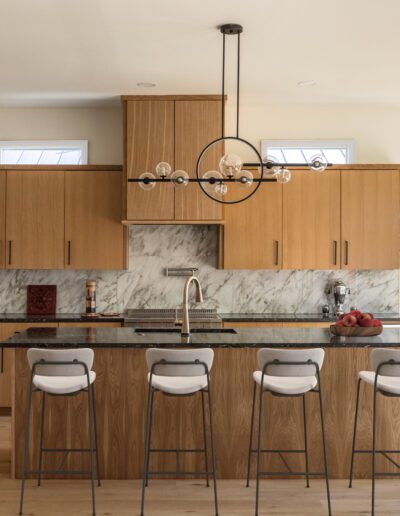 Modern kitchen with light wood cabinets, marble backsplash, black countertop, island with four stools, and a contemporary chandelier above. Large window provides natural light.
