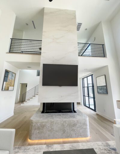 A modern living room with a tall marble fireplace, wall-mounted TV, light wood floors, large windows, and contemporary artwork.
