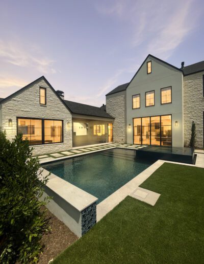 Modern two-story house with large windows, stone exterior, and illuminated interiors, featuring a rectangular swimming pool and landscaped lawn at dusk.