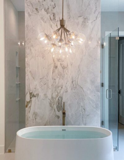A modern bathroom with a freestanding oval bathtub, marble accent wall, glass shower enclosure, and a contemporary chandelier overhead.