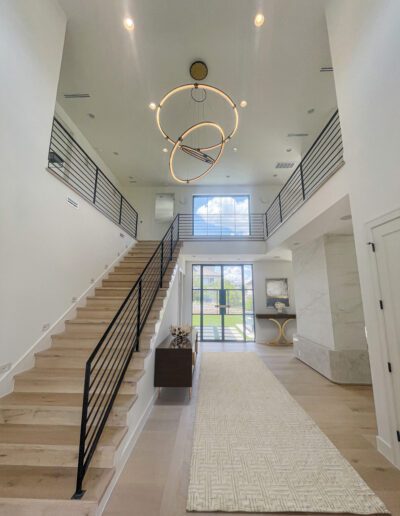 Modern, spacious foyer with light wood floors, a large geometric chandelier, open staircase with black railings, and a floor-to-ceiling window letting in natural light.