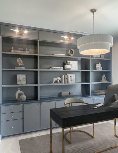 A modern home office with built-in dark shelves, a black desk with gold legs, a computer, a chair, decorative objects, books, and a round overhead pendant light.