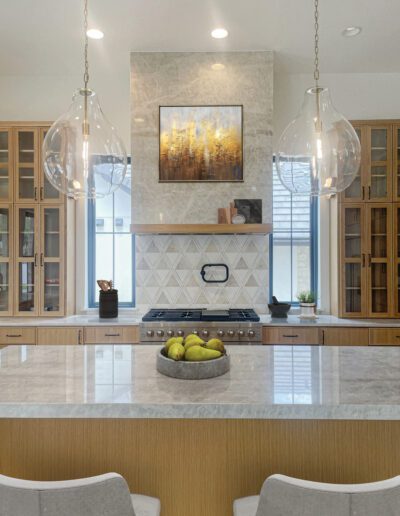 Modern kitchen with marble island, wooden cabinets, pendant lights, a bowl of green apples, and a stove beneath a decorative geometric backsplash. Framed art hangs above the range.
