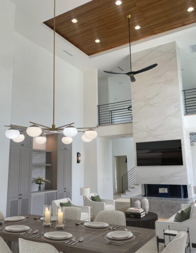 Modern open-concept living and dining area with high ceilings, marble fireplace, dining table set for six, contemporary chandelier, and neutral-toned decor.