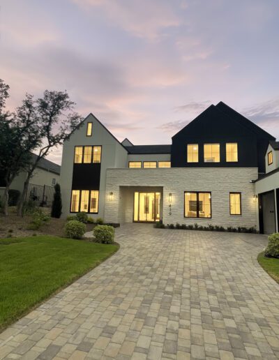 Modern two-story house with large windows, a brick and black exterior, a lit entryway, and a paved driveway at dusk.