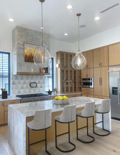 Modern kitchen with wood cabinets, marble island and countertops, four barstools, pendant lights, stainless steel appliances, and a fruit bowl with lemons on the island.