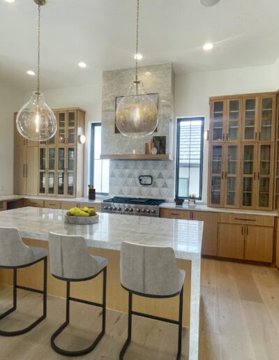Modern kitchen with light wood cabinets, a large marble island with four chairs, stainless steel appliances, and pendant lights above the island.