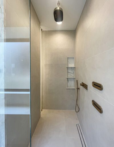 Modern, minimal walk-in shower with beige tiles, built-in shelves, a handheld showerhead, two horizontal grab bars, and a ceiling light.