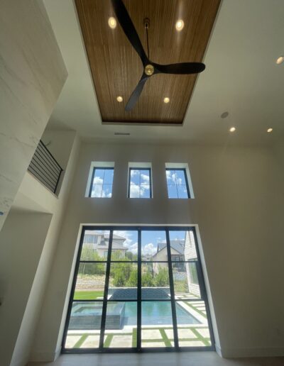 View from inside a modern living room with high ceiling, ceiling fan, large windows, and a backyard pool visible outside.
