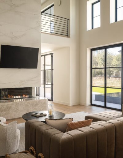 A modern living room with tall windows, a brown sectional sofa, two white chairs, a marble fireplace with a mounted TV, and a staircase with metal railing.