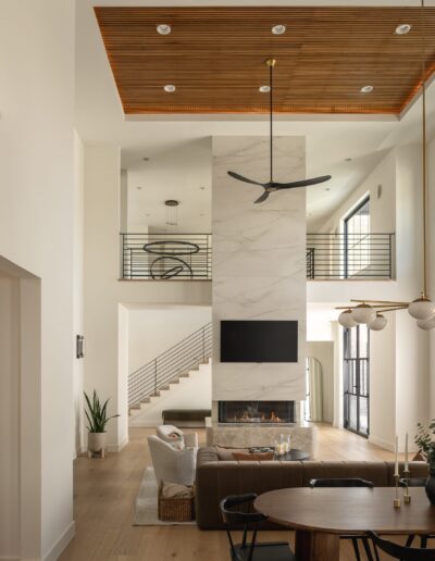 Modern living room with high ceilings, a marble fireplace, wall-mounted TV, hanging ceiling fan, wooden accents, large windows, and a staircase leading to an open upper floor.