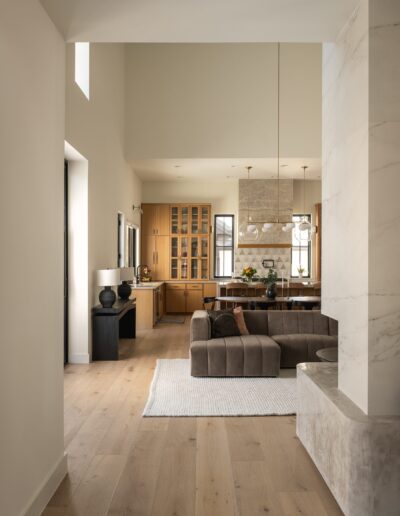 Modern living room with light wood flooring, a brown sofa, white rug, stone accent wall, and a view into a kitchen with wooden cabinets and a dining table.