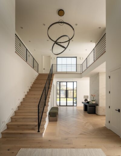Modern entryway with light wood floors, open staircase with black railings, a large geometric chandelier, and floor-to-ceiling windows letting in natural light.