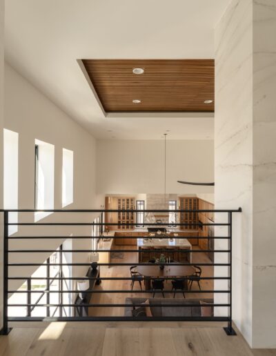 Modern open-concept living and dining area viewed from an upper floor, featuring wood flooring, large windows, and a suspended light fixture.