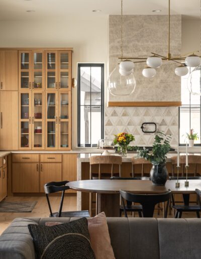Modern kitchen with wooden cabinets, glass-paneled doors, pendant lights, a large island with stools, and a dining table with chairs in an open-plan living space.