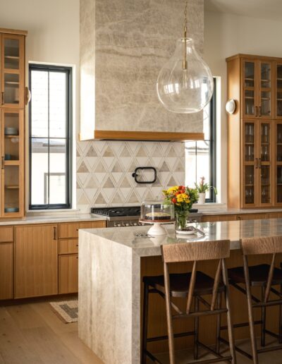 Modern kitchen with wooden cabinets, marble island and backsplash, three bar stools, a glass pendant light, flowers on the counter, and daylight coming through two narrow windows.