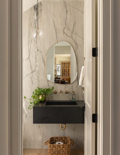 Modern bathroom with a black floating sink, oval mirror, marble wall, hanging light, potted plant, towel ring, and woven basket on light wood floor.