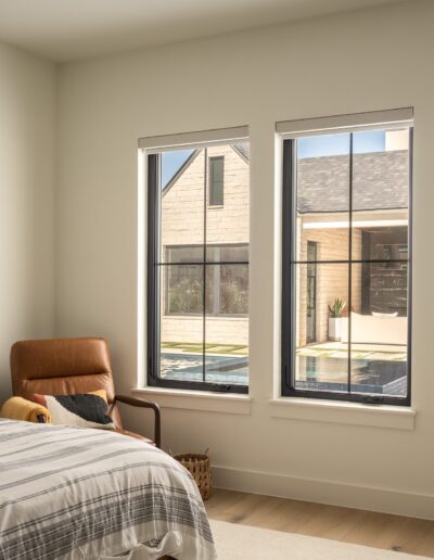 A modern bedroom with a bed, brown leather chair, and three large windows overlooking a pool and patio area outside.