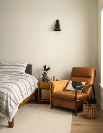 A bedroom with a bed, striped bedding, a wooden nightstand, a potted plant, a brown leather armchair, throw pillows, a basket, and a wall-mounted lamp.
