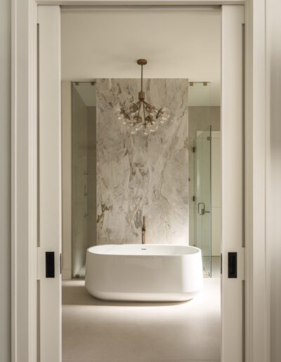 Modern bathroom with a white freestanding bathtub, marble accent wall, glass shower enclosure, and a contemporary chandelier overhead.