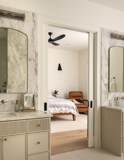 Modern bathroom with two vanities, marble countertops, and arched mirrors; sliding door opens to a bedroom with a brown chair and bed.