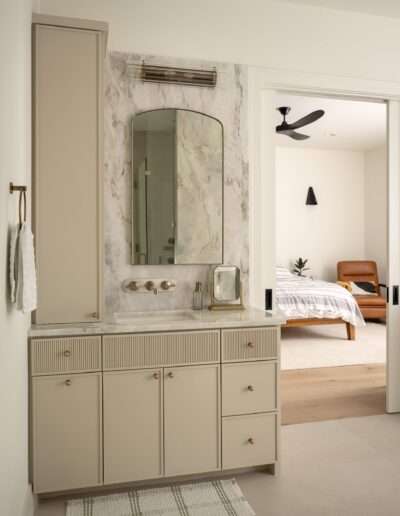 Modern bathroom vanity with beige cabinets, marble backsplash, rectangular mirror, wall-mounted faucet, and view into a bedroom with a wooden bed and ceiling fan.