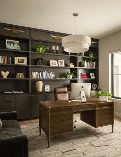 Modern home office with dark built-in bookshelves, a wooden desk with an iMac, indoor plants, and a large window letting in natural light.