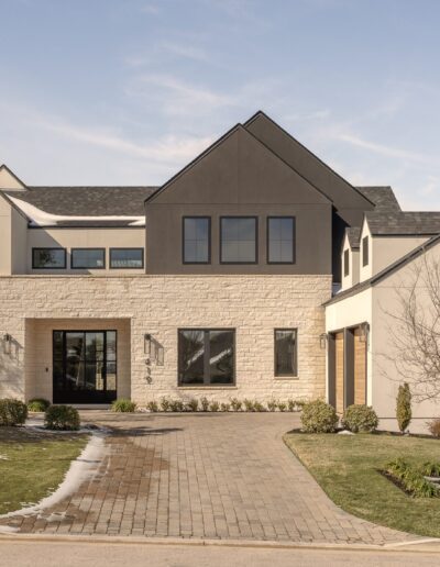 Modern two-story house with a mix of stone and stucco exterior, large windows, and a paved driveway, surrounded by a manicured lawn and minimal landscaping.