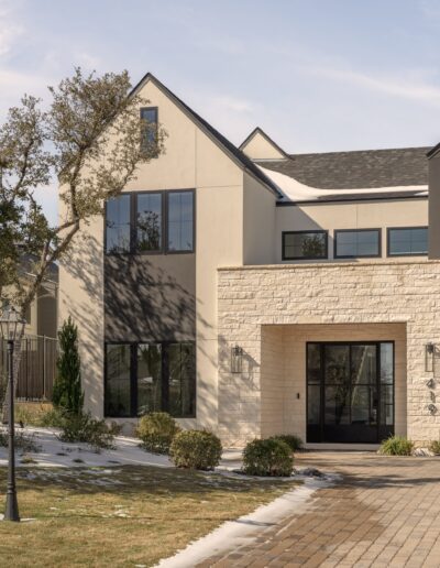 Modern two-story house with cream-colored walls, large windows, stone accents, and a front entrance. Light snow and shadows are visible on the ground and roof.