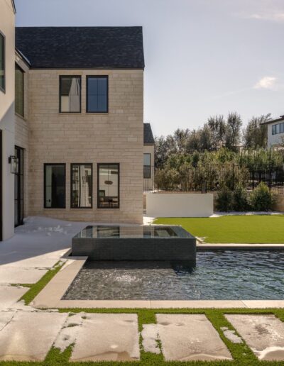 Modern house with large windows overlooks a backyard featuring a pool, attached hot tub, artificial grass, and stone pavers, with neighboring houses visible in the background.