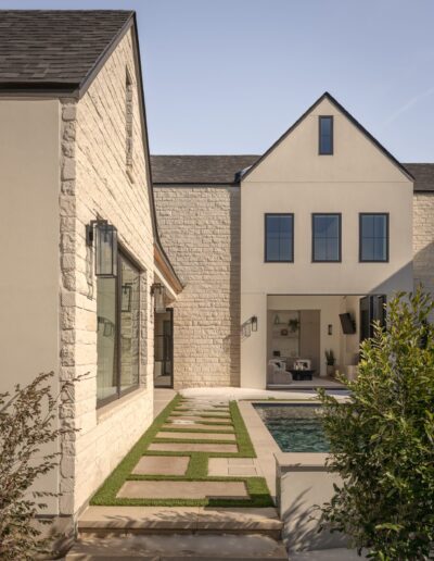 Modern two-story house with stone and stucco exterior, large windows, and a small poolside patio, viewed from a walkway lined with grass and concrete pavers.