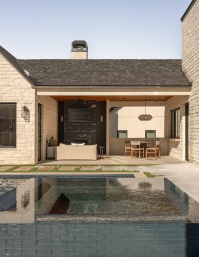 Modern house exterior with stone walls, a pool and hot tub in the foreground, covered patio area with dining table, and large windows and doors.
