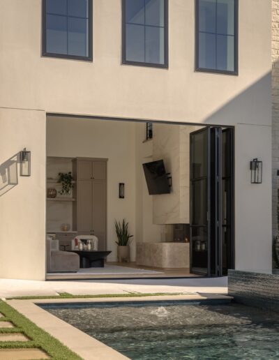 Modern house with large open glass doors leads from a living room to an outdoor patio and pool. The interior features neutral colors and minimalist decor.